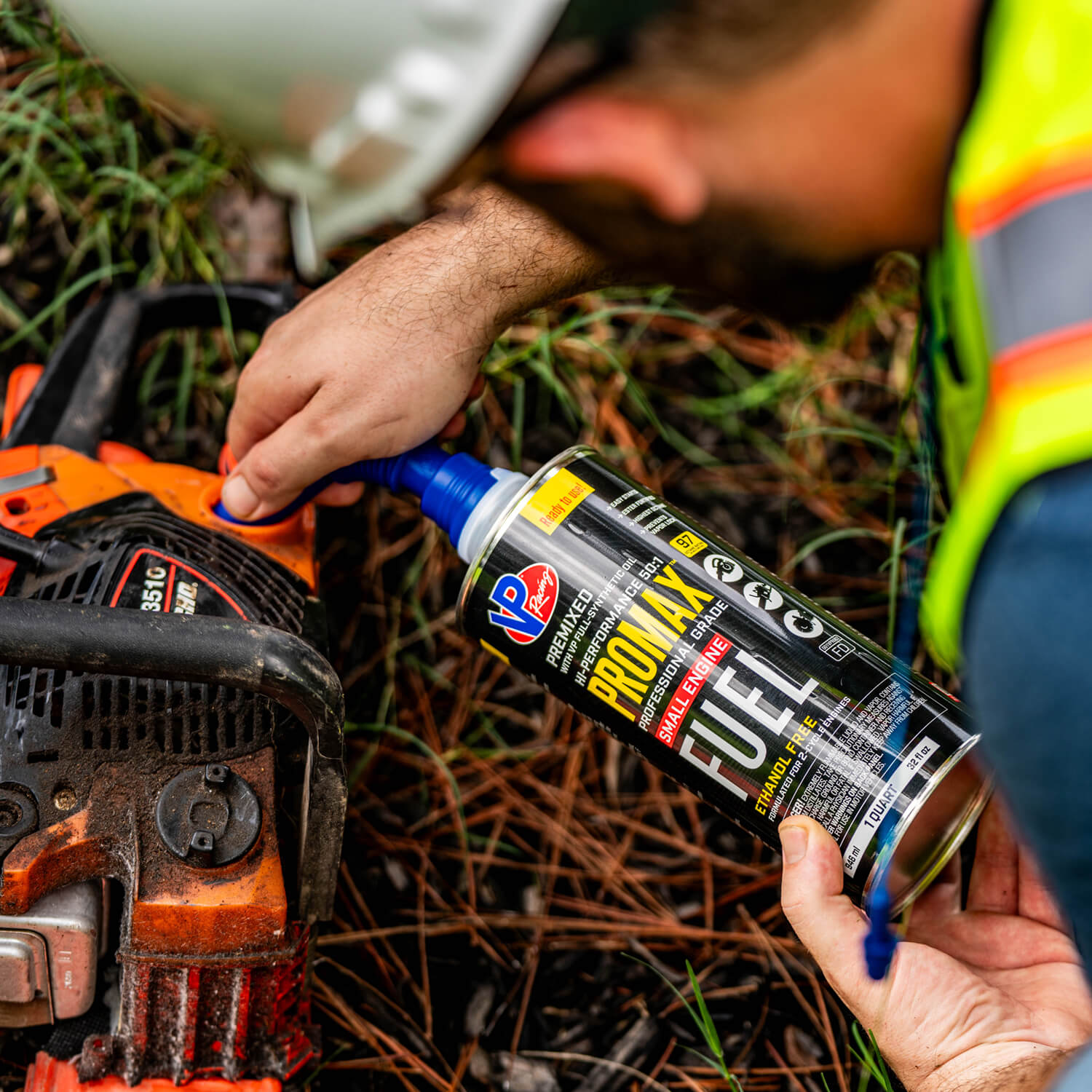 Worker in safety vest adding VP ProMax fuel to an orange chainsaw engine, surrounded by grass.