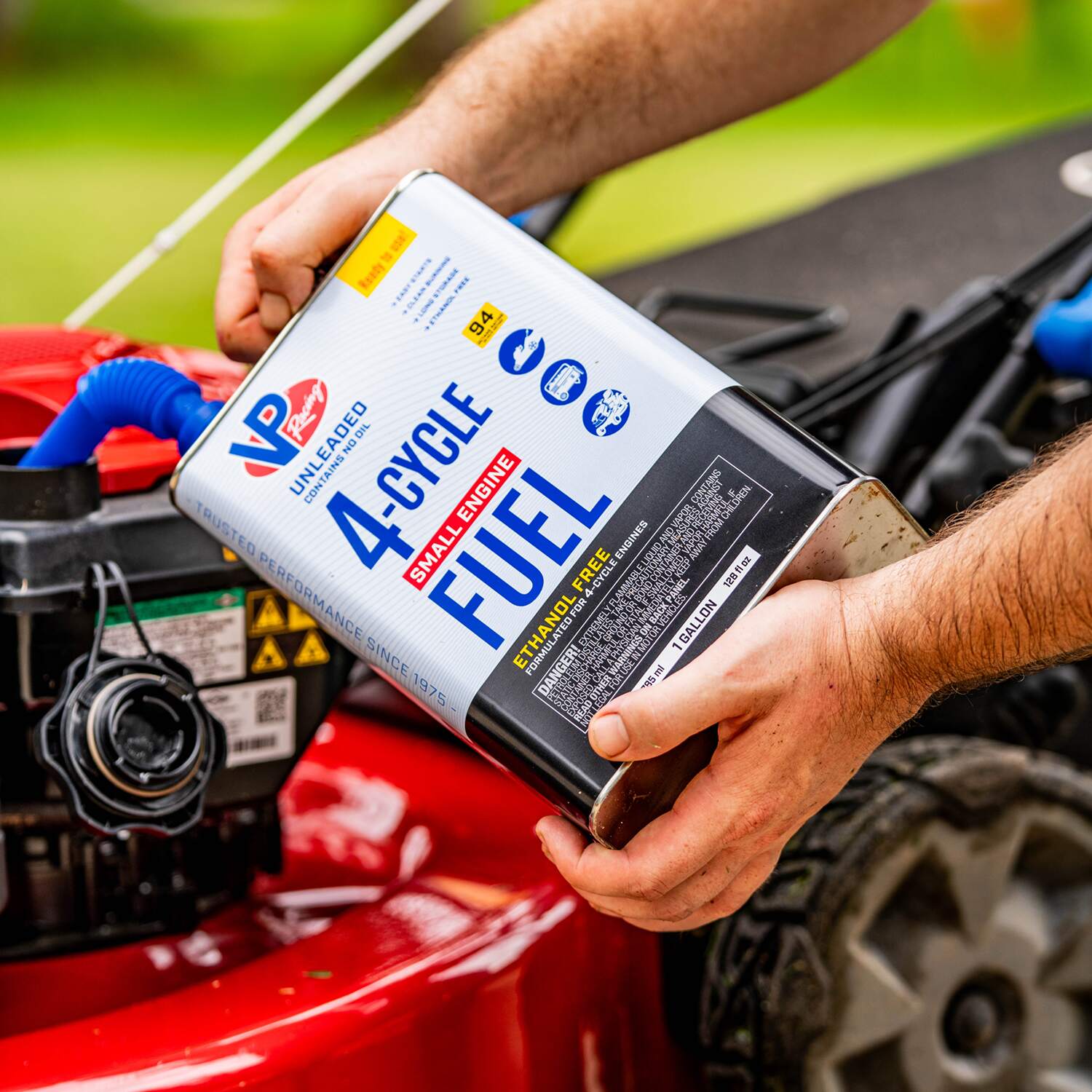 A person pouring VP Racing 4-Cycle fuel from a one-gallon container into the small engine of a red lawn mower.