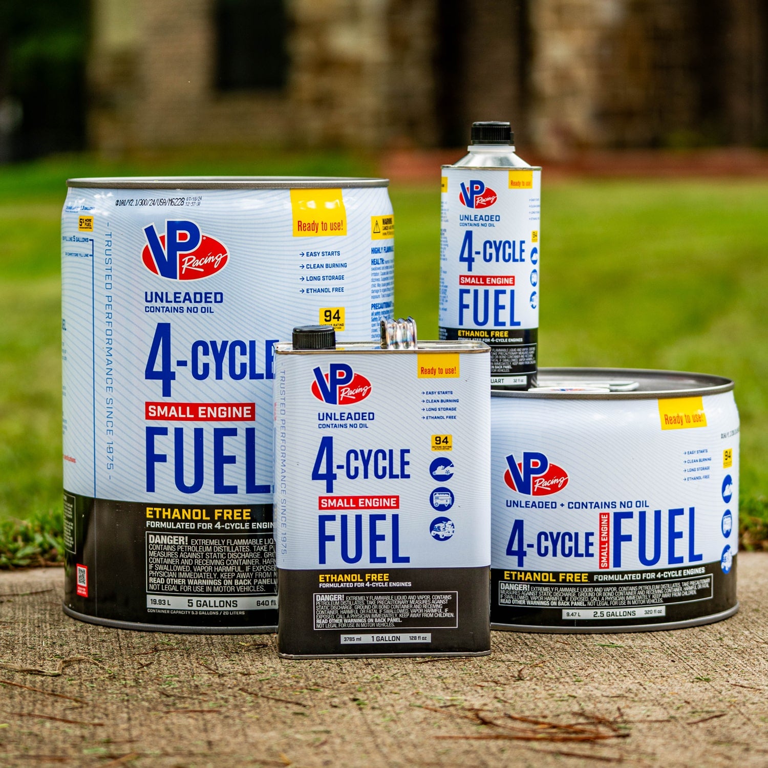 Different container sizes of VP Racing 4-Cycle Fuel for small engines. The containers are arranged outdoors on concrete, ranging from quarts to large pails.