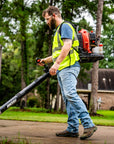 A landscaping worker in safety vest using backpack leaf blower on path, with lawn and trees behind him.