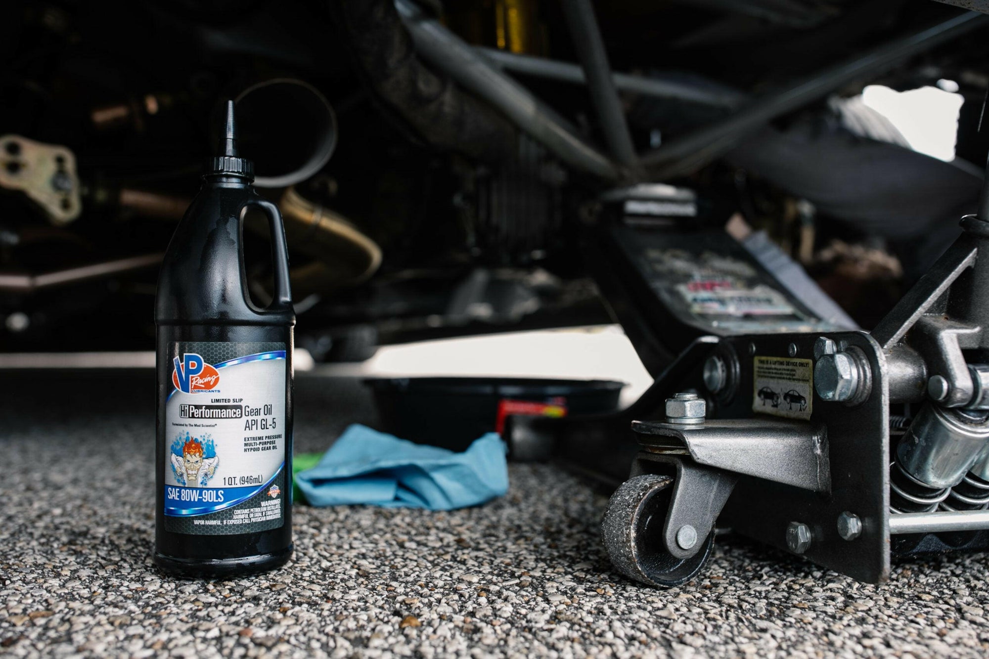 lifestyle image of a bottle of VP Hi-Performance 80w90 GL5 gear oil sitting on the ground beneath a car that has been raised on a jack