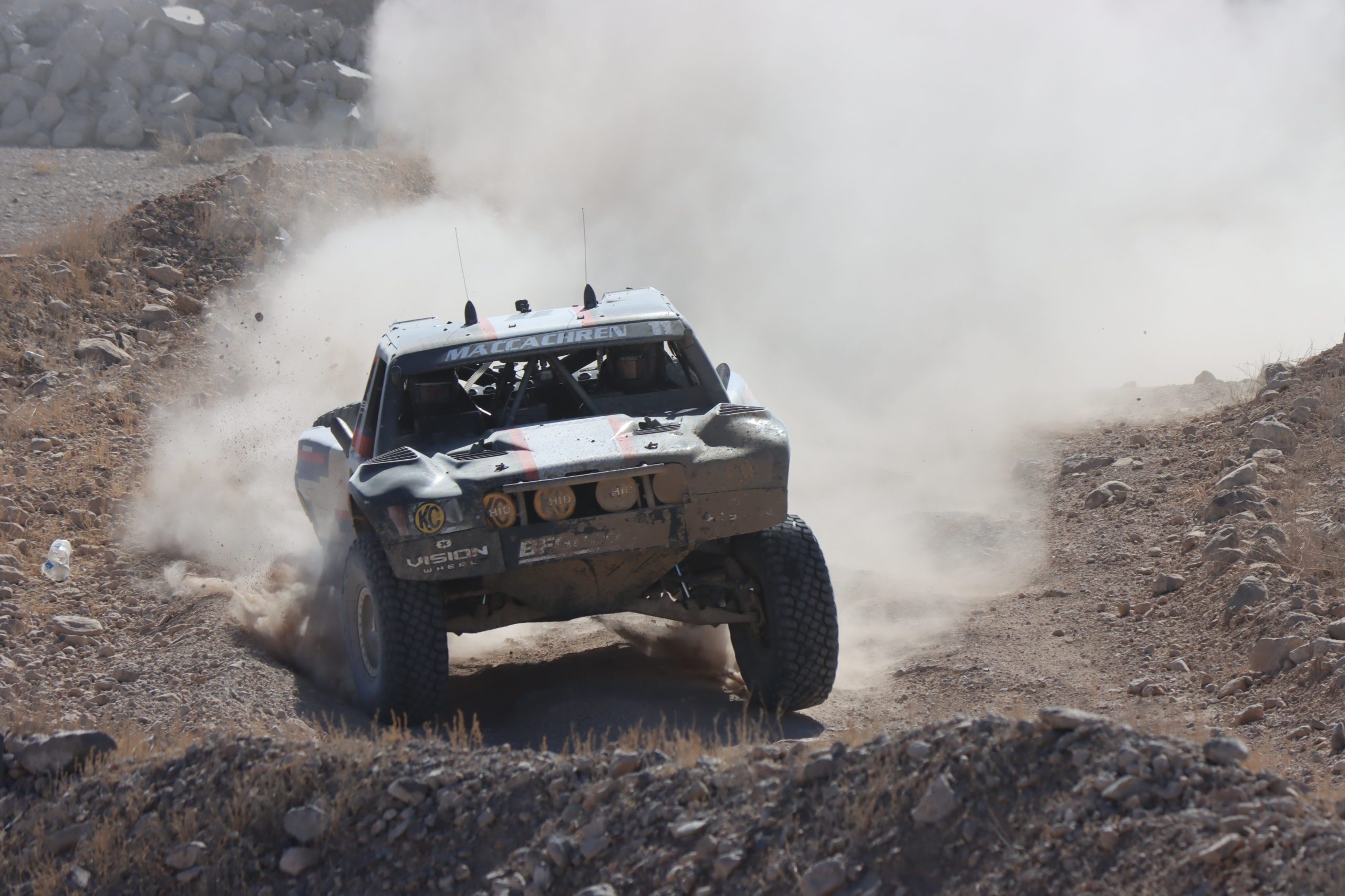 action photo of Rob MacCachren and his trophy truck racing through the desert in the afternoon. A huge dust cloud of dirt is trailing his truck, which is coming toward the camera