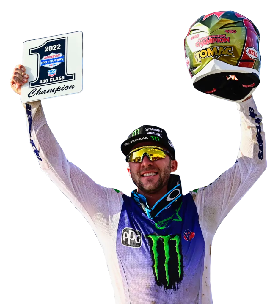 Eli Tomac holding his arms up with his motorcycle helmet in his left hand and a sign in his right hand that shows he won 1st place in the 450 class motocross