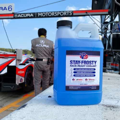 64 ounce bottle of Stay Frosty Race-Ready engine coolant. Bottle is on a concrete ledge in the pit stop area of a race track. There is a race car in the background.