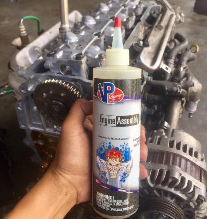 Bottle of VP engine assembly lube being held in the hand of an unknown person. There is an engine on the floor in the immediate background.