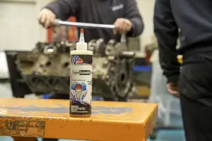 A bottle of VP Assembly Lube on a work bench. An engine is on a table in the background with an unrecognizable man working on it